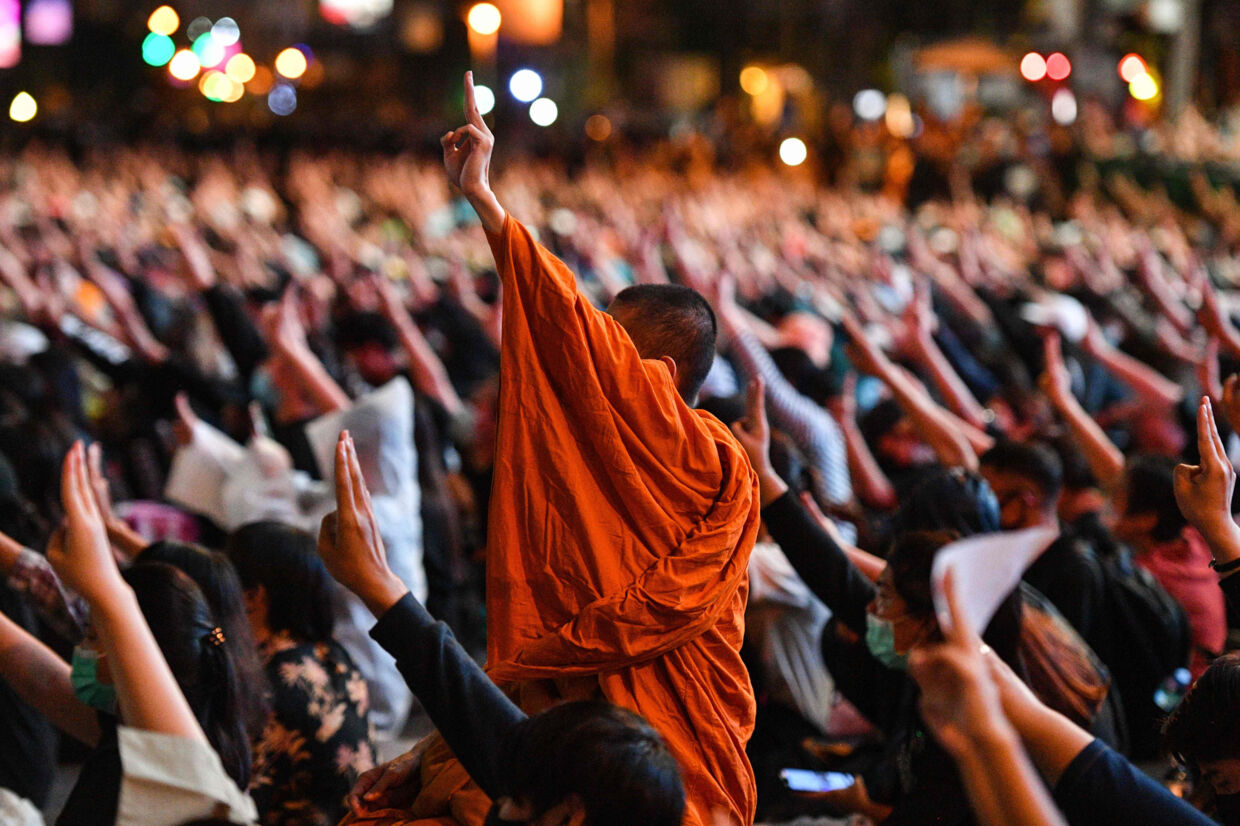 En buddhistmunk og tusinder af andre demonstranter løfter deres hænder og viser tre fingre - den hilsen, der er blevet demokratibevægelsens kendetegn - under søndagens demonstration i Bangkok. Mladen Antonov/Ritzau Scanpix