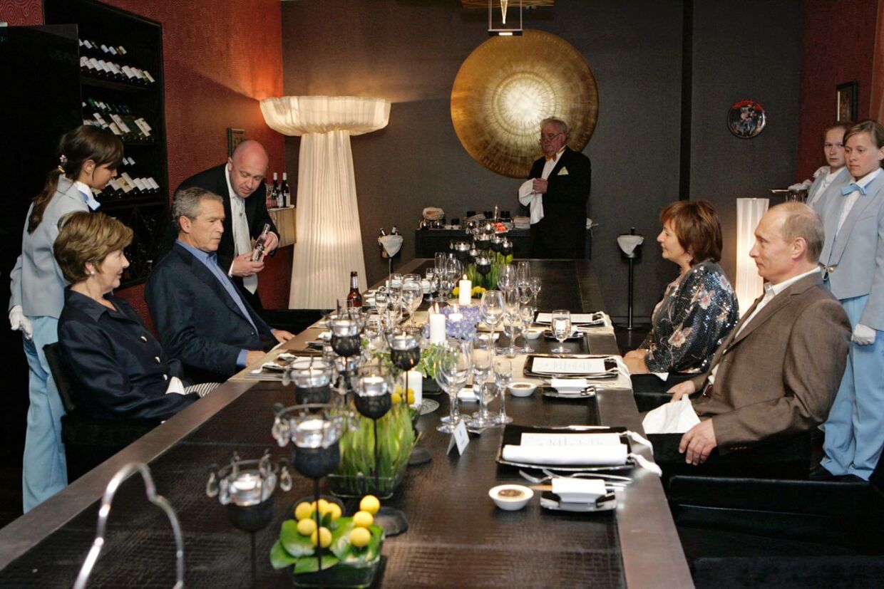 På højre side af bordet ses Vladimir Putin og den daværende hustru, Ljudmila Putina. På venstre side af bordet ses George og Laura Bush – bagest ses Jevgenij Prigozjin.